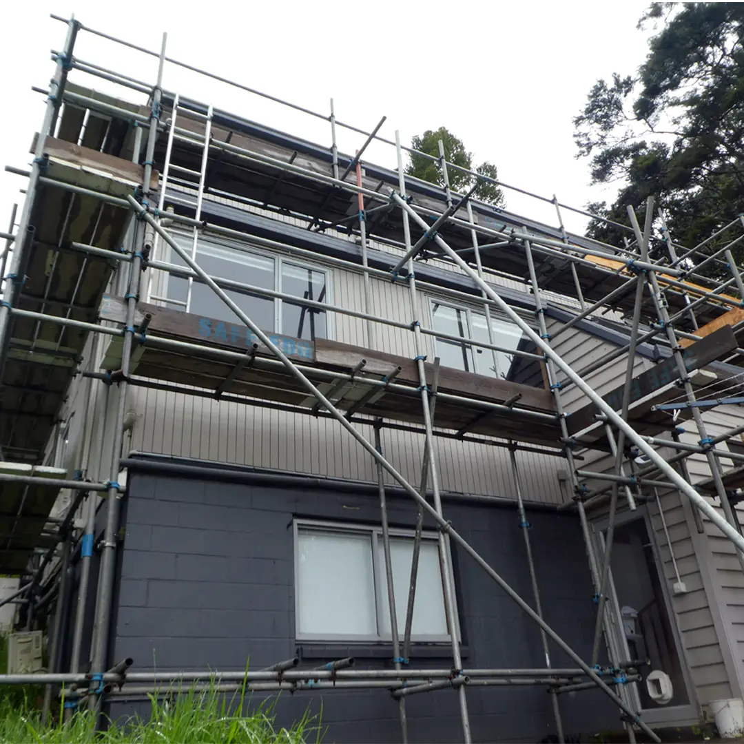 The image is of a house being painted with scaffolding around it, set against a backdrop of the sky and trees. The house is in the process of exterior painting.