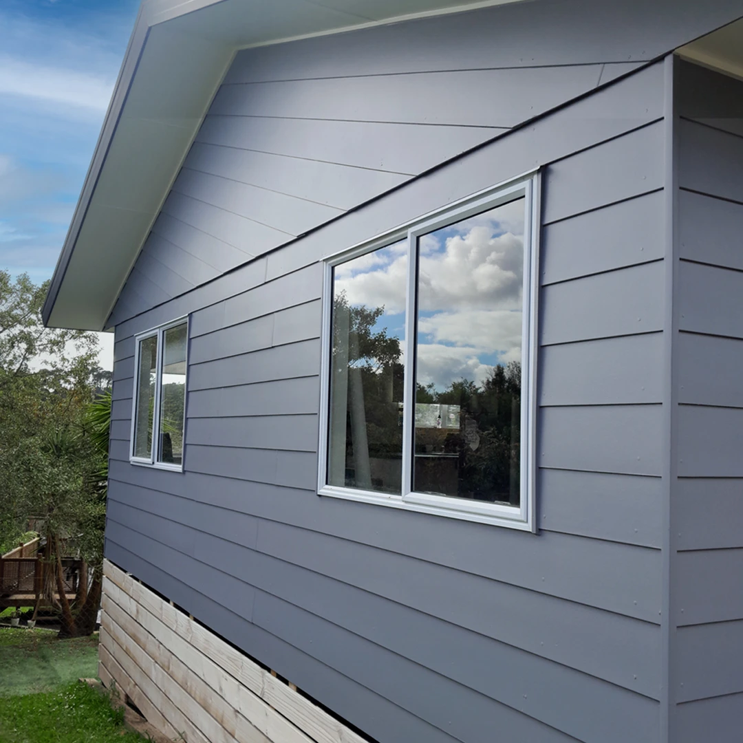 Gray house exterior with two rectangular windows reflecting clouds, set against a blue sky. White trim and wooden foundation visible. Trees and greenery in the background.