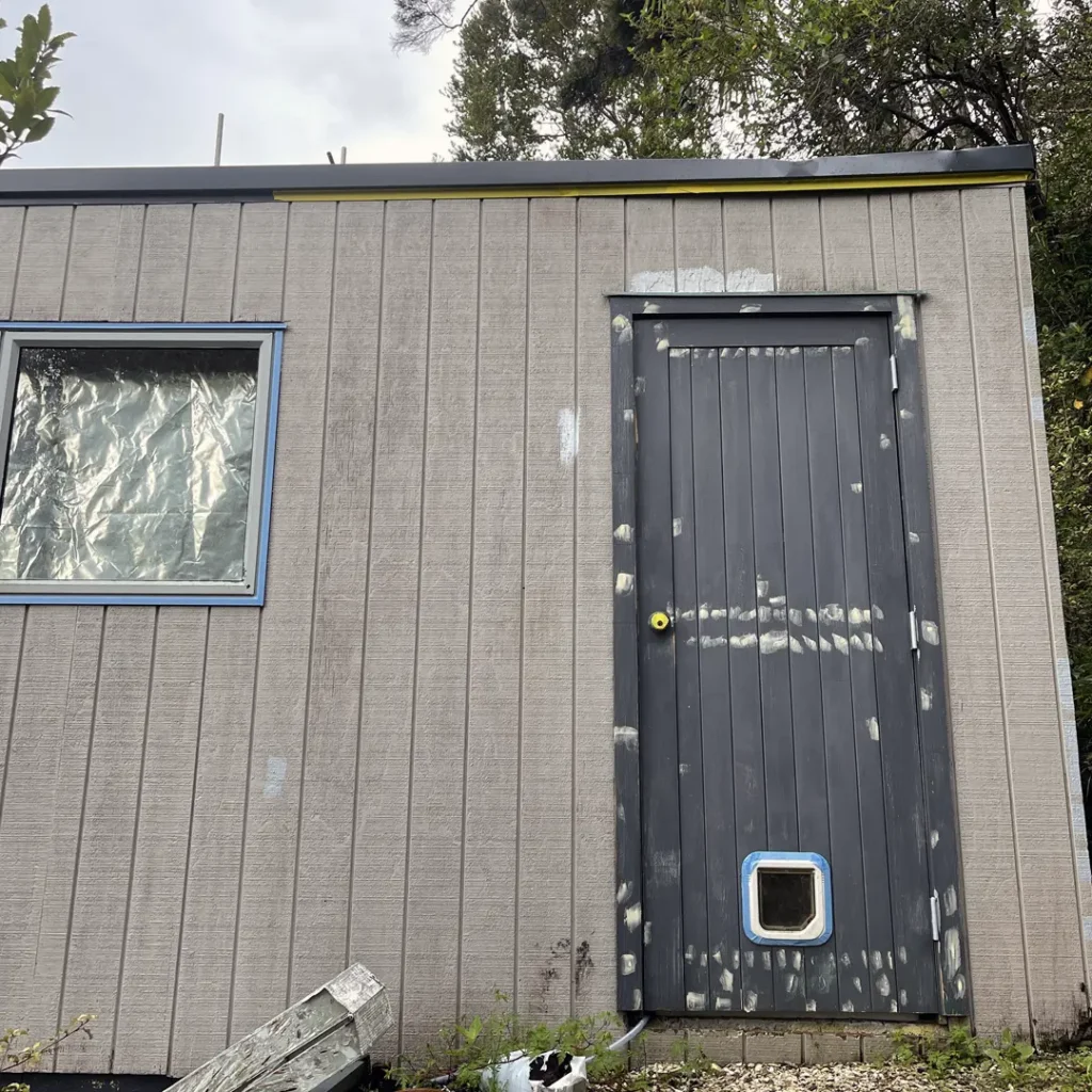 The image shows a house exterior before painting, reflecting visible signs of aging and neglect. The walls are faded and discolored, with patches of peeling paint and uneven surfaces. The trim and window frames appear weathered and chipped, lacking definition and a polished look. The front door blends into the overall dull appearance, offering no standout feature. The overall condition of the exterior suggests the need for thorough preparation and a fresh coat of paint to restore its appeal and protect it from further wear.