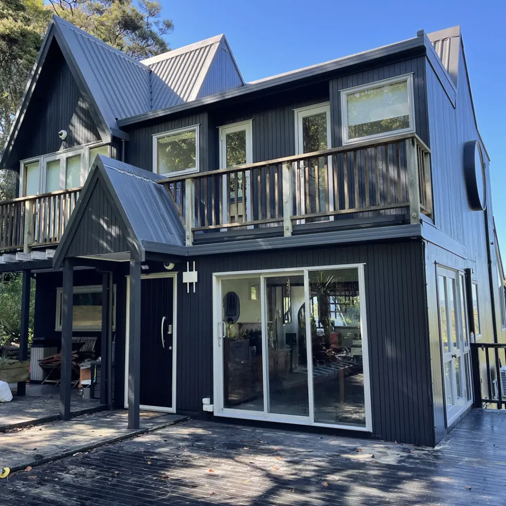 The image depicts a dark house showcasing the exterior after being painted with Weathershield® ColourGuard™ Rawene. It features elements such as aluminium windows, a deck, a round window and vertical cladding.